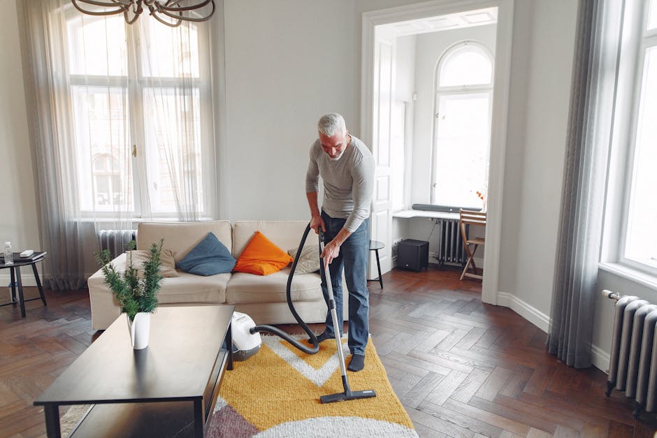 A person wearing beige shoes, blue jeans, and a beige coat is vacuuming a patterned area rug with a handheld vacuum cleaner in a room with soft lighting; the rug features floral and geometric motifs i